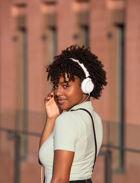 Attraente ragazza latino-americana dai capelli ricci che ascolta musica con le cuffie bianche per strada. Dal retro guardando la telecamera - Foto, immagini