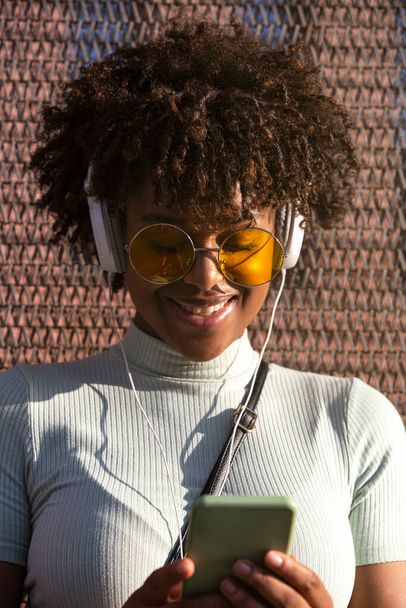 Cool joven latinoamericana con cabello afro y gafas sonrientes usando smartphone para redes sociales. - Foto, Imagen