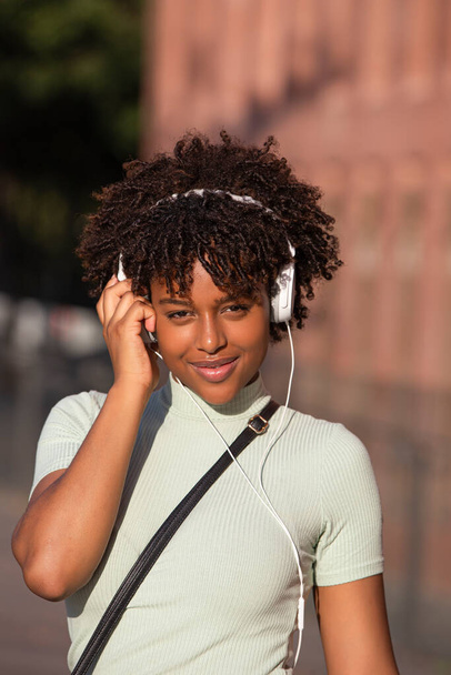 Cheerful smiling Latin American woman with curly hair listening to music on smartphone wears earphones in the street, looking at camera. - 写真・画像