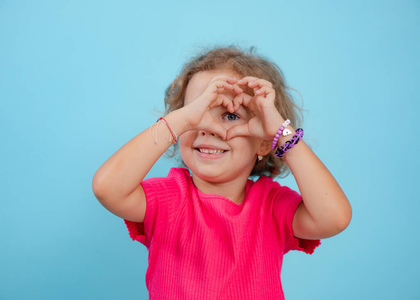 Portrait of happy blue-eyed little girl with curly fair hair wearing pink T-shirt, looking through fingers making heart shape on blue background. Studio. Advertisement, emotions, feelings, copy space. - Photo, image
