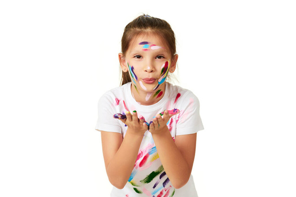 happy little child girl sending an air kiss with hands painted in colorful paint isolated on white background. - 写真・画像