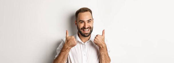 Close-up van tevreden man met baard in wit shirt, met duimen omhoog in goedkeuring, als en eens, positief antwoord, witte achtergrond. - Foto, afbeelding