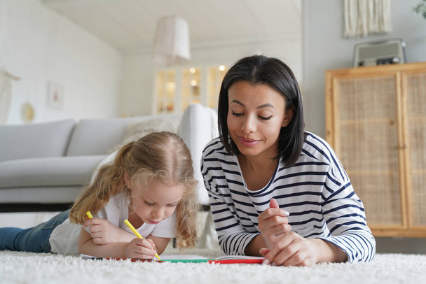 Μητέρα ή μπέιμπι-σίτερ και υιοθετημένη κόρη ζωγραφίζουν μαζί ξαπλωμένη στο χαλί του πατώματος στο σπίτι, μαμά με το κορίτσι προσχολικής ηλικίας ζωγραφίζοντας με πολύχρωμα μολύβια. Ανάπτυξη παιδικής δημιουργικότητας, έννοια της υιοθεσίας. - Φωτογραφία, εικόνα