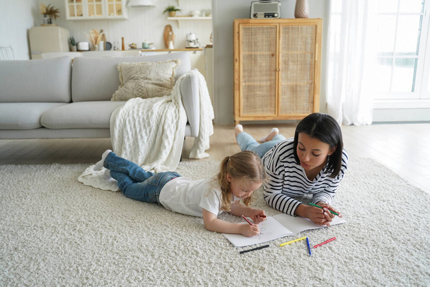 Mom or babysitter and little preschooler kid girl painting together lying on floor carpet in living room. Mother and daughter drawing. Child creativity development, children's education concept. - Photo, Image