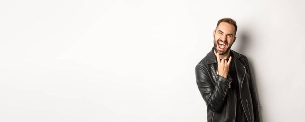 Cool adult man in black leather jacket, showing rock on gesture and tongue, enjoying music festival, standing over white background. - Foto, Bild