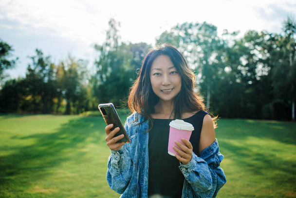 happy asian girl smiling and using mobile phone while walking in summer park at sunset. - Photo, image