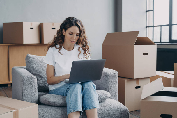 Focused hispanic girl works at laptop searching ideas for renovation, choosing furniture, shopping interior decor for new house online, sitting on armchair among card boxes. Moving, relocation concept - Photo, Image
