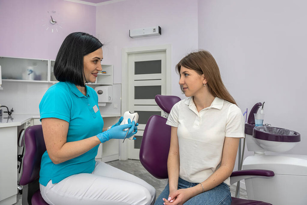 Πορτρέτο ενός ευτυχισμένου ασθενή με υγιή δόντια κάθεται σε μια οδοντιατρική καρέκλα δίπλα σε μια νεαρή γυναίκα οδοντίατρος. εξέταση του ασθενούς. Διαβούλευση σε οδοντιατρική κλινική - Φωτογραφία, εικόνα