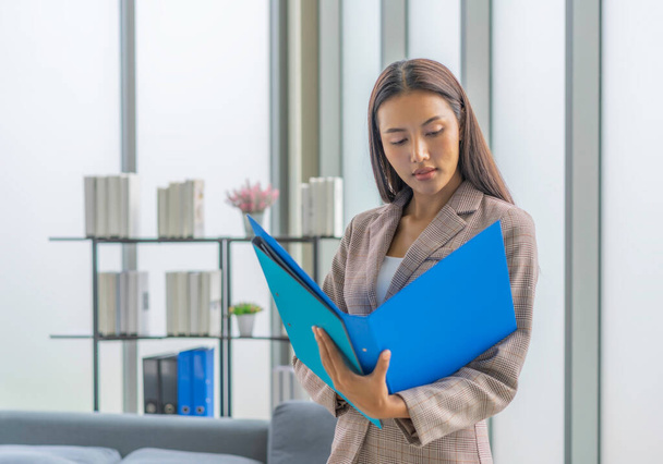 Крупным планом красотка азиатская бизнесвумен стоит у окна в рабочее время и вдумчиво анализирует отчет в синем файле. - Фото, изображение