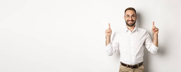 Confident handsome businessman showing logo, pointing and looking up at announcement, standing over white background. - Photo, Image