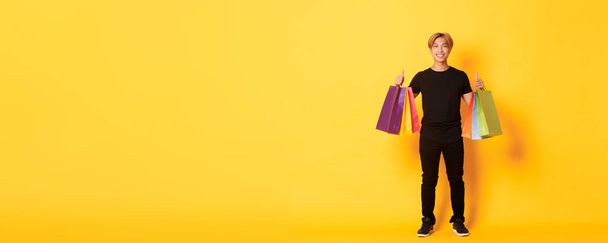 Полноразмерный привлекательный корейский парень в черной одежде, показывающий большие пальцы вверх, держащий сумки с покупками, желтый фон. - Фото, изображение