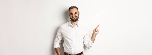 Skeptical male entrepreneur grimacing, pointing finger left with displeased face, complaining on product, white background. - Photo, Image