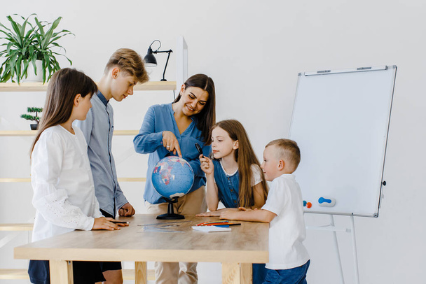 Teacher and children in class are looking at globe, teacher helps explain the lesson to the children in the class at a desk. Educational school process, bright room and interesting learning - 写真・画像