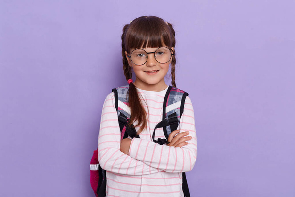 Horizontal shot of pretty positive little girl with pigtails wearing backpack standing isolated over purple background, posing with folded hands, looking at camera with smile. - Fotografie, Obrázek