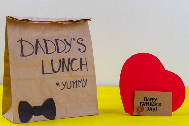 Paper bag and red heart on yellow wooden desk. Daddys lunch for fathers day. - Photo, Image