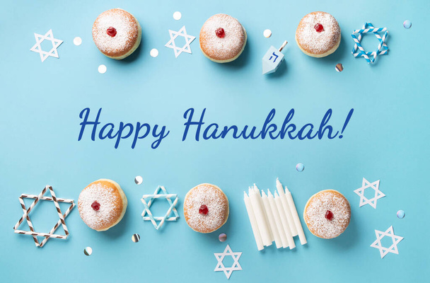 Hanukkah sweet doughnuts sufganiyot (traditional donuts) with fruit jelly jam, gift boxes, spinnig driedel and candles on blue paper background. Jewish holiday Hanukkah concept. Top view, copy space. - 写真・画像