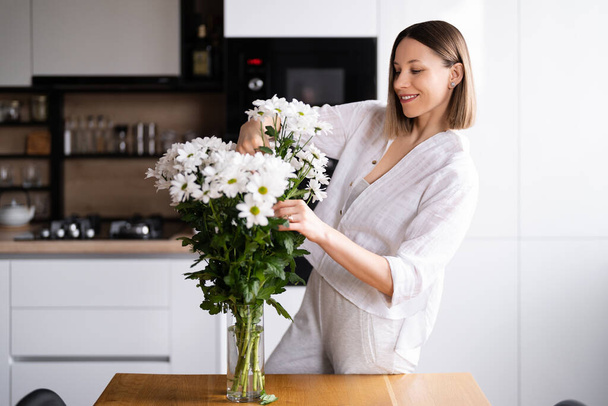 Jovem mulher feliz e alegre em branco organizando flores brancas em casa na cozinha - Foto, Imagem