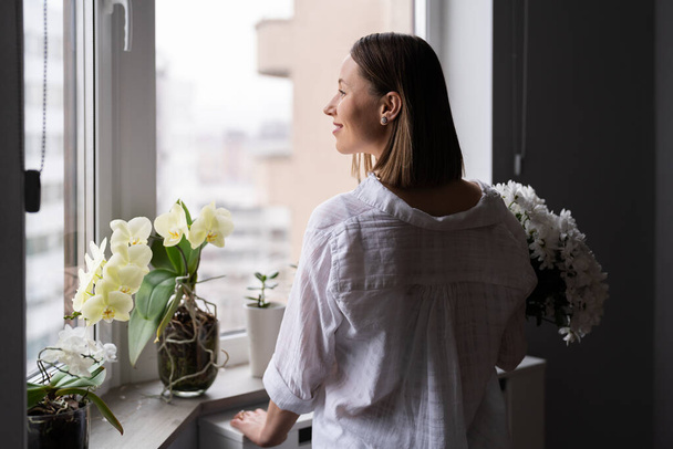 Jeune femme portant des vêtements blancs décontractés regardant par la fenêtre tenant un bouquet de fleurs blanches attendant le printemps ou l'été à venir - Photo, image