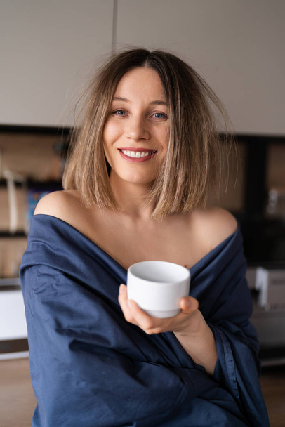 Álmos, pozitív nő, kék lepedőbe csomagolva, kávét iszik reggel a konyhában. - Fotó, kép