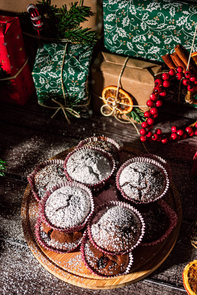 Powdered sugar muffins on wooden plate and Christmas gifts. New year boxes with kraft, red, green paper decoration. Still life close up. Food photo. - Photo, image