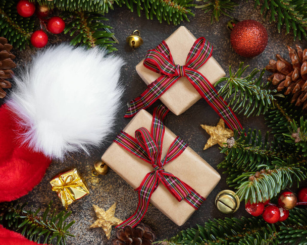Завернутые подарочные коробки между рождественскими украшениями, шляпа Санта-Клауса и зеленые ели ветви вид сверху на темном черном фоне. Рождественская или новогодняя атмосфера - Фото, изображение