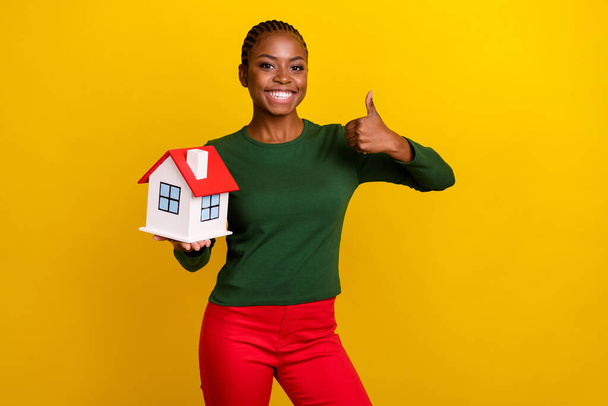Photo of shiny confident lady dressed green shirt thumb up holding small house isolated yellow color background. - Photo, Image