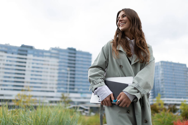 Porträt einer jungen Studentin mit Laptop und Buch, die vor dem Hintergrund städtischer Gebäude lächelnd in die Ferne blickt. - Foto, Bild