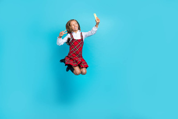 Full size portrait of small beautiful schoolgirl jump hold telephone make selfie show v-sing isolated on blue color background. - Foto, Imagen