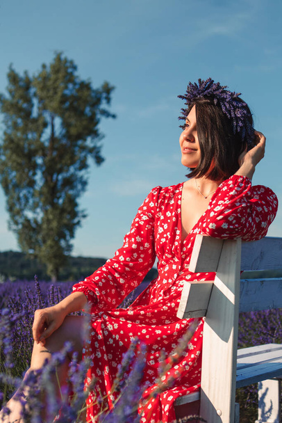 una giovane donna con un vestito rosso e una corona sulla testa siede su una panchina bianca in un campo di lavanda - Foto, immagini