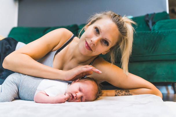 Znaczenie snu w życiu niemowlęcia. Średnie zbliżenie wewnątrz ujęcie pięknej białej matki pochylającej się nad niemowlęciem, które śpi na szarym materacu umieszczonym na podłodze, oraz - Zdjęcie, obraz