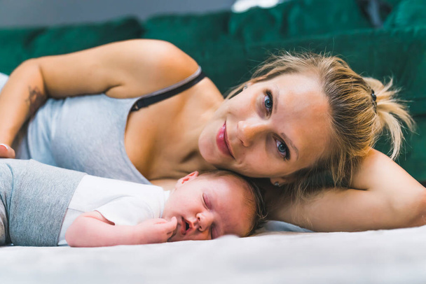 Closeup indoor portrait of a middle-aged blond caucasian mom in gray tank top looking and smiling at camera while leaning over her adorable sleeping baby infant boy. High quality photo - Photo, Image