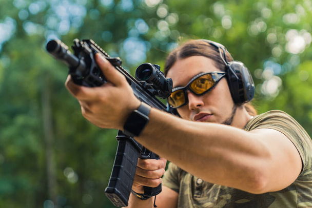  Hombre caucásico con gafas protectoras y auriculares practicando con un rifle negro en el campo de tiro, primer plano medio. Foto de alta calidad - Foto, Imagen