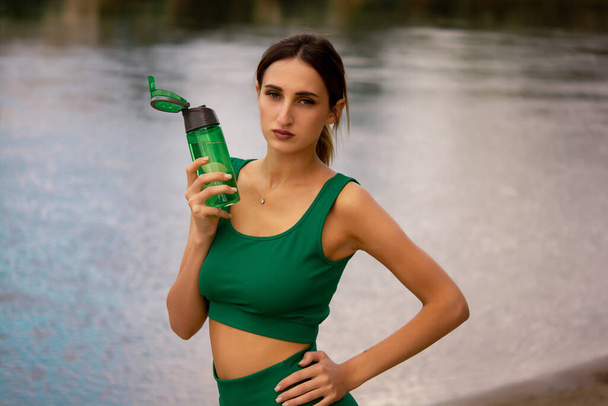 egy lány sport egyenruhában egy üveget tart a kezében, egy lány egy üvegből iszik vizet, egy lány zöld melegítőbe van öltözve, és rövidnadrágban biciklizik, vízegyensúly, szabadtéri sportok, sport életmód - Fotó, kép