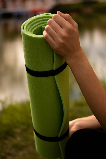 Egy lány sportegyenruhában jóga szőnyeget tart a kezében, fekete sportfelsőben, zöld szőnyegben, jóga a parkban, sportéletmód, szabadtéri sportok. - Fotó, kép