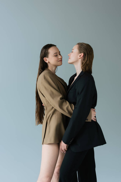 pretty woman with long hair embracing lesbian girlfriend in black blazer isolated on grey - Foto, afbeelding