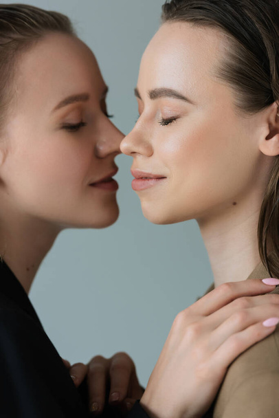 side view of smiling lesbian girlfriends standing face to face with closed eyes isolated on grey - Foto, imagen