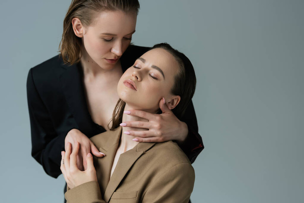 morena mujer con los ojos cerrados cerca joven lesbiana novia abrazando su aislado en gris - Foto, Imagen