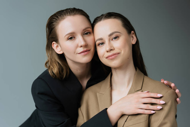 young and pleased lesbian partners in beige and black jackets looking at camera isolated on grey - Foto, Bild