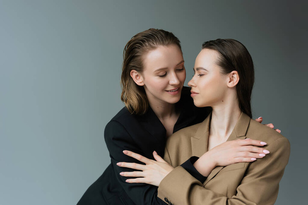 smiling lesbians in beige and black blazers hugging isolated on grey - Foto, Imagem