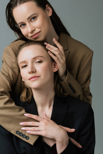 young and pleased lesbian partners in blazers looking at camera and embracing isolated on grey - 写真・画像
