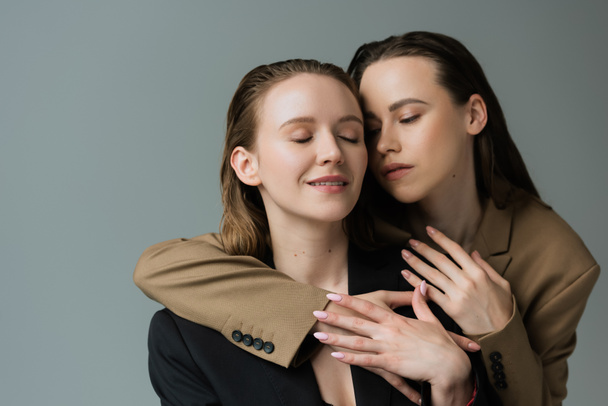 brunette woman in beige blazer embracing lesbian girlfriend smiling with closed eyes isolated on grey - Photo, Image
