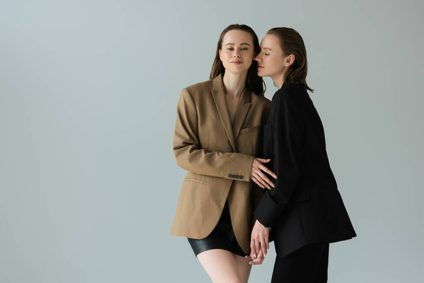 pretty lesbian woman in black suit holding hands with girlfriend in beige blazer isolated on grey - Фото, изображение