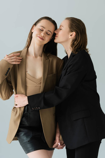 mladá žena v černé sako objímající přítelkyně stojící se zavřenýma očima izolované na šedé - Fotografie, Obrázek