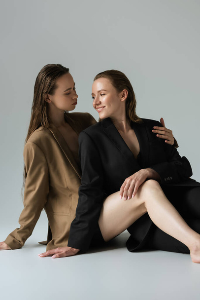 same sex lesbian couple in blazers smiling at each other while sitting on grey - Foto, Imagem