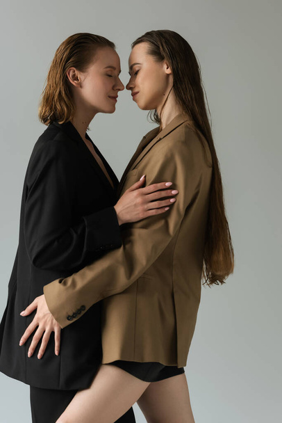Seitenansicht lesbischer Frauen in beigen und schwarzen Blazern, die mit geschlossenen Augen und isoliert auf grau stehen - Foto, Bild