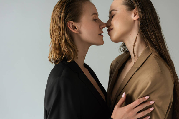 boční pohled na lesbické ženy se zavřenýma očima stojící tváří v tvář izolované na šedé - Fotografie, Obrázek
