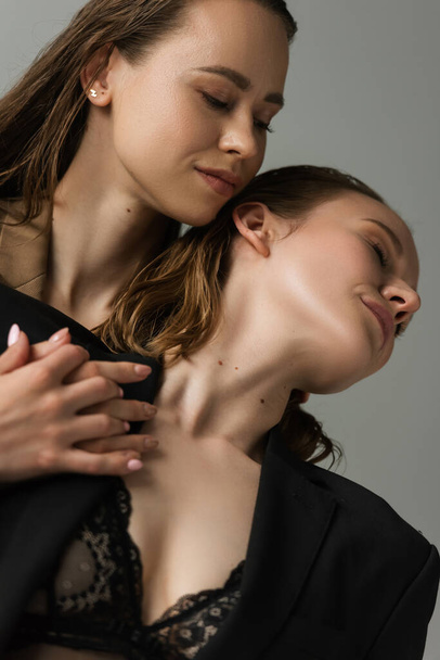low angle view of young lesbian woman near seductive girlfriend in black bra and jacket isolated on grey - Foto, Imagen