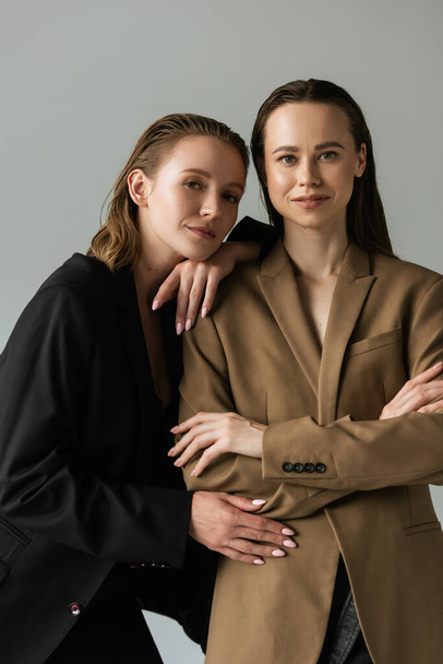 young woman leaning on lesbian girlfriend standing with crossed arms and looking at camera isolated on grey - Fotografie, Obrázek