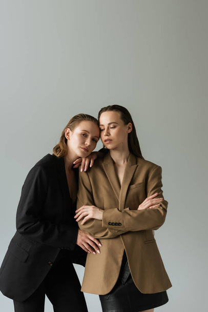 lesbian woman in beige blazer standing with crossed arms near pretty girlfriend isolated on grey - Фото, изображение