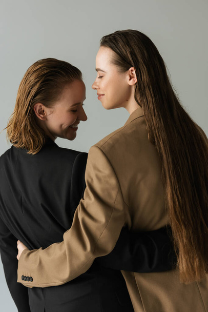 Rückansicht von lesbischem Paar in schwarzen und beigen Blazern, die vereinzelt auf grau lächeln und sich umarmen - Foto, Bild